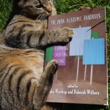 A tabby cat plays with a copy of the para-academic handbook on the grass.